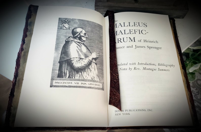 May include: An open book with a brown leather cover and a bookmark. The book is titled "The Malleus Maleficarum" by Heinrich Kramer and James Sprenger. The book is translated with introductions, bibliography, and notes by Rev. Montague Summers. Dover Publications, Inc. New York.