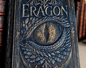 May include: A hardcover book titled Eragon with a dark blue cover featuring an embossed dragon eye design. The eye is surrounded by textured scales and intricate gold-toned filigree patterns, with the title text prominently displayed at the top.