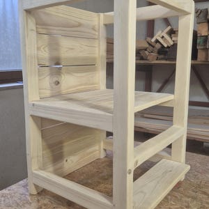 May include: A natural wood learning tower with a square frame. The tower has a platform, a step, and a back panel made of horizontal wooden slats. The tower is designed to help children reach countertops and other elevated surfaces.
