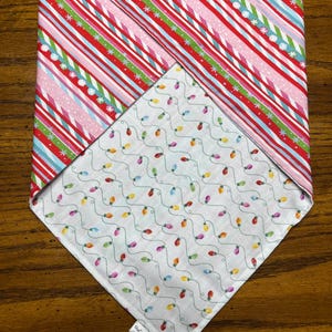 May include: A triangular pet bandana featuring a double-sided design. One side displays diagonal stripes in red, pink, light blue, and green with white snowflake accents. The reverse side features a white background with colorful string lights.