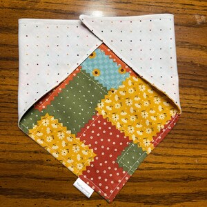 Peut inclure: Un bandana triangulaire pour animal avec un motif patchwork de fleurs et de formes géométriques dans des tons jaune moutarde, vert olive et rouge terre cuite. Le revers est en tissu blanc à petits points multicolores avec une étiquette.