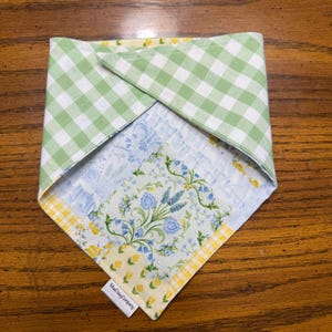 Può includere: Un bandana reversibile per animali domestici. Un lato presenta un motivo a quadri verdi e bianchi, l'altro un design floreale con fiori blu e dettagli gialli. L'etichetta recita "MadDaisyCompany".