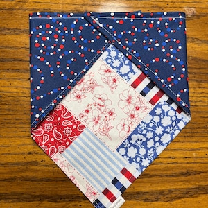 May include: A patriotic dog bandana with a patchwork design. The bandana features a navy blue background with red and white polka dots, and various patterned squares including paisley, floral, and striped patterns in red, white, and blue. The tag reads "Mad Dawg Company".