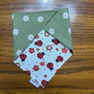May include: A square pet bandana with a green daisy print on one side and a white background with red ladybugs and flowers on the other. The bandana is folded in half, showing both patterns. The tag reads "MadDogCompany".