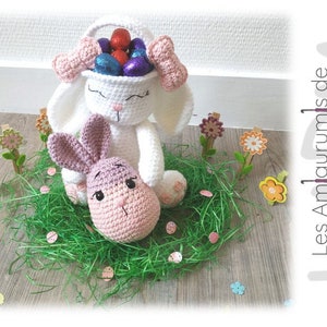 May include: A white crocheted bunny holding a basket filled with colorful Easter eggs. The bunny is sitting on a bed of green grass with pink and white Easter eggs scattered around it.  The bunny is holding a smaller pink crocheted bunny.