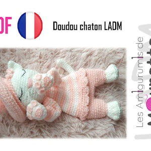 May include: A white crocheted cat with pink stripes, wearing a pink and white crocheted hat. The cat is lying on a pink fuzzy surface. The text "Doudou chaton LADM" is visible in the image.