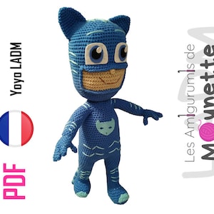 May include: A blue crocheted amigurumi toy of a superhero character wearing a mask. The character has a cat-like face with large eyes and a smile. The toy is standing on a white background.