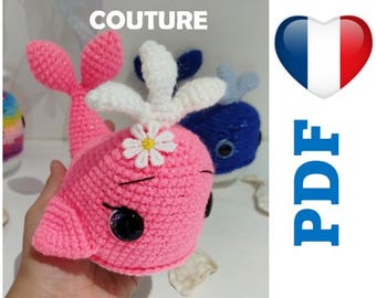 May include: A pink crocheted whale with a white flower on its head. The whale has black eyes and a white crocheted tail. The image also includes the text "SANS COUTURE" and "PDF" in blue.