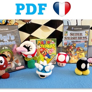 May include: Crocheted Super Mario Bros. characters, including a Goomba, a Piranha Plant, a Koopa Troopa, a mushroom, and a Bob-omb. The characters are displayed on a blue surface with video game cases in the background.