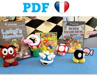 May include: Crocheted Super Mario Bros. characters, including a Goomba, a Piranha Plant, a Koopa Troopa, a mushroom, and a Bob-omb. The characters are displayed on a blue surface with video game cases in the background.
