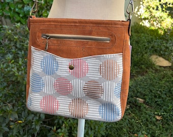 May include: A brown leather crossbody bag with a zippered pocket and a front flap. The flap has a white fabric lining with a pattern of blue, pink, and brown circles on a white background.