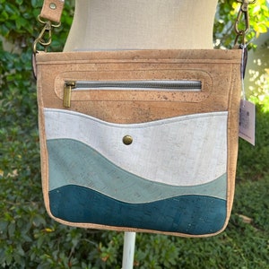 Puede incluir: Un bolso bandolera de corcho y tela con un patrón de ondas en tonos azules, blancos y beige. El bolso tiene un bolsillo con cremallera en la parte delantera y una correa larga ajustable.