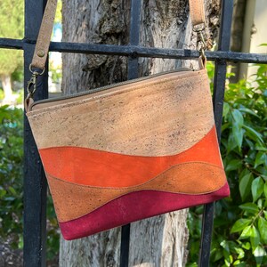 Puede incluir: Un bolso bandolera de corcho con cierre de cremallera. El bolso tiene un dise&ntilde;o ondulado con tres rayas horizontales en tonos naranja, marr&oacute;n y burdeos.