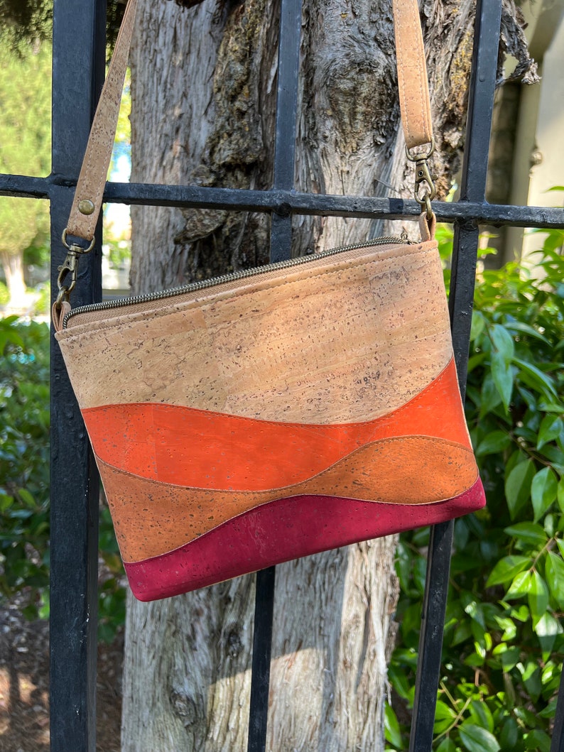Puede incluir: Un bolso bandolera de corcho con cierre de cremallera. El bolso tiene un dise&ntilde;o ondulado con tres rayas horizontales en tonos naranja, marr&oacute;n y burdeos.