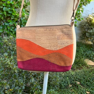Puede incluir: Un peque&ntilde;o bolso bandolera de corcho con cierre de cremallera. El bolso tiene un dise&ntilde;o ondulado en tres colores: naranja, marr&oacute;n y burdeos.