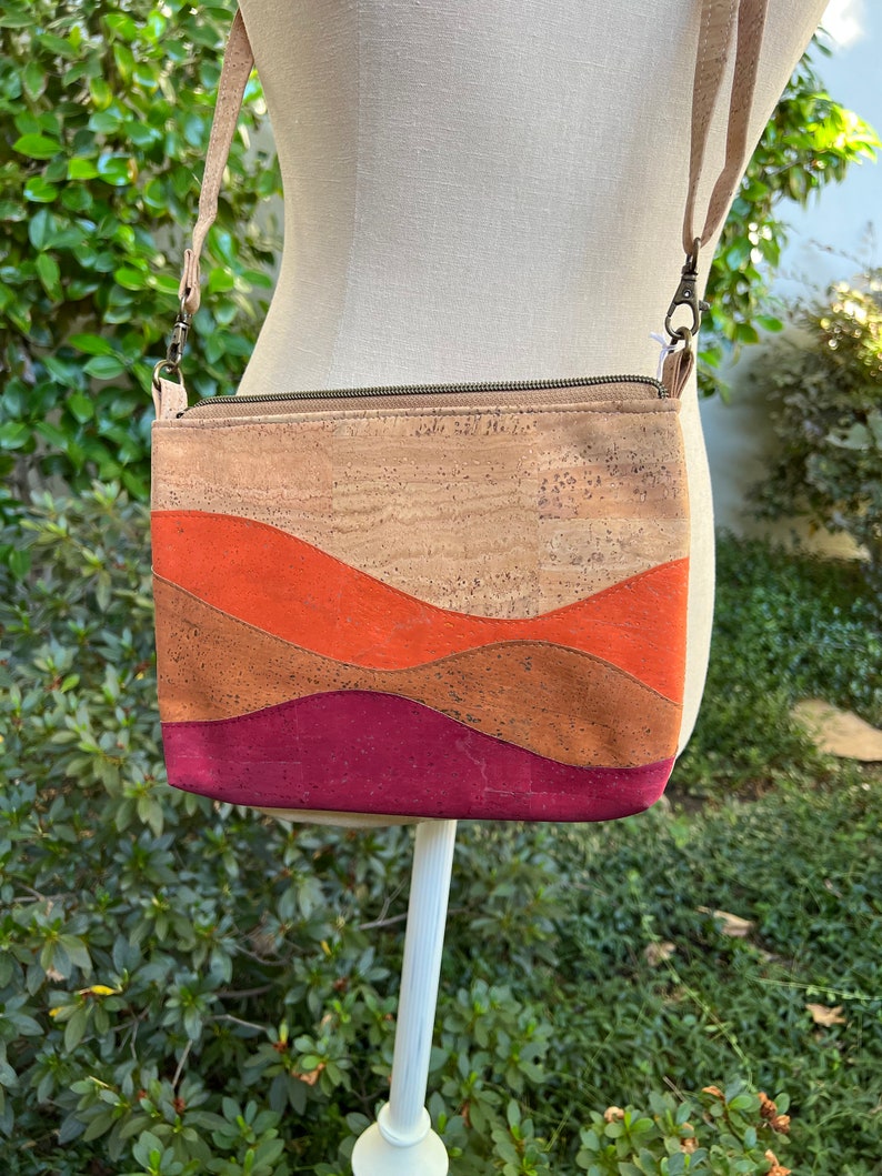 Puede incluir: Un peque&ntilde;o bolso bandolera de corcho con cierre de cremallera. El bolso tiene un dise&ntilde;o ondulado en tres colores: naranja, marr&oacute;n y burdeos.