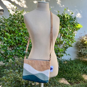 Puede incluir: Un peque&ntilde;o bolso bandolera de corcho con un dise&ntilde;o ondulado en tonos azules, blancos y marrones. El bolso tiene una cremallera y una larga correa ajustable.