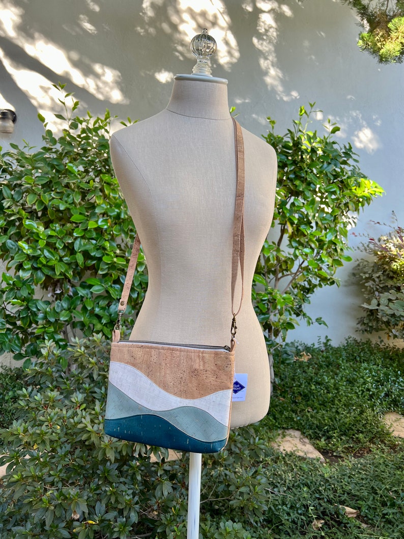 Puede incluir: Un peque&ntilde;o bolso bandolera de corcho con un dise&ntilde;o ondulado en tonos azules, blancos y marrones. El bolso tiene una cremallera y una larga correa ajustable.