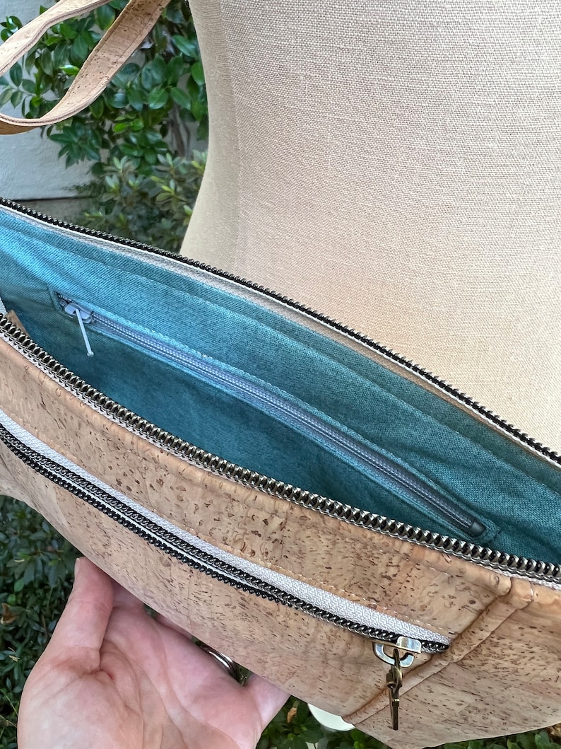 Puede incluir: Un bolso de mano de corcho con cierre de cremallera y forro de tela turquesa. El bolso tiene un asa superior y un bolsillo con cremallera en la parte delantera.