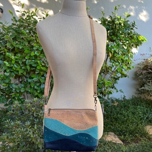 Puede incluir: Un peque&ntilde;o bolso bandolera de corcho con un patr&oacute;n ondulado azul, turquesa y marr&oacute;n. El bolso tiene una correa larga ajustable y un cierre de cremallera.