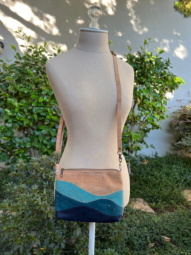 Puede incluir: Un peque&ntilde;o bolso bandolera de corcho con un patr&oacute;n ondulado azul, turquesa y marr&oacute;n. El bolso tiene una correa larga ajustable y un cierre de cremallera.