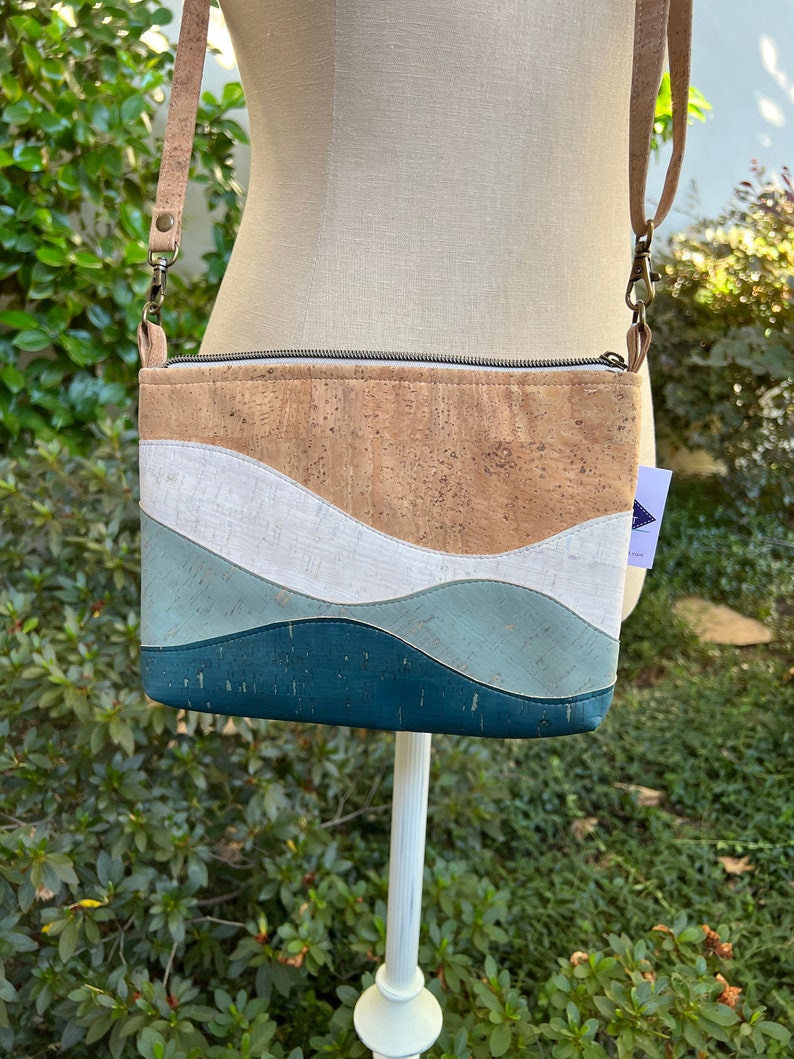 Puede incluir: Un peque&ntilde;o bolso bandolera de corcho y cuero con cierre de cremallera. El bolso tiene un dise&ntilde;o ondulado en tonos azules, blancos y marrones.