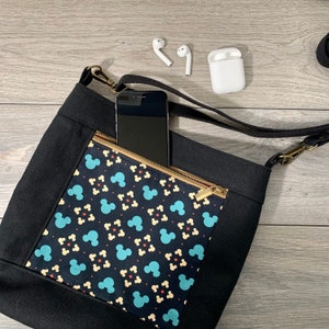 May include: A black canvas crossbody bag with a zippered pocket featuring a blue and yellow Mickey Mouse print. A black smartphone is in the pocket.