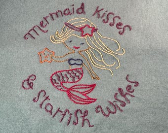 May include: A teal fabric with embroidered text and a mermaid design. The text reads "Mermaid Kisses & Starfish Wishes." The mermaid has blonde hair, a red starfish, and a red tail. The embroidery is in shades of red, gold, and purple.