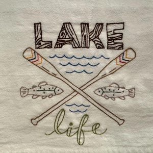 May include: Embroidered white towel with the words "LAKE" in brown, wood-grain style letters. Two crossed oars, blue water waves, two fish, and the word "life" in green script complete the design. A lake-themed item.