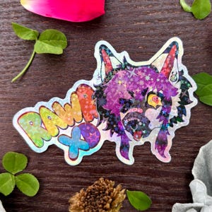 May include: Holographic sticker featuring a cartoon wolf with purple and black fur, adorned with stars. The sticker has the text "RAW XP" in rainbow colors. The sticker is on a wooden surface with leaves and a flower petal.