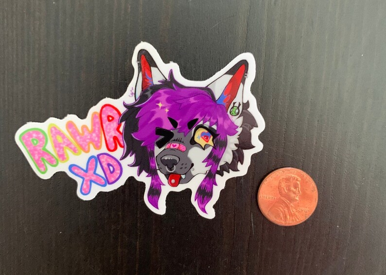 May include: A colorful sticker featuring a cartoon wolf with purple and black fur, a winking eye, and a tongue sticking out. The sticker has the words "RAWR XD" in a rainbow font. A penny is next to the sticker for size comparison.