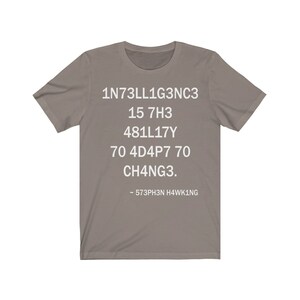 May include: A gray t-shirt with white text that reads "1N73LL1G3NC3 15 7H3 481LL17Y 70 4D4P7 70 CH4NG3. - 573PH3N H4WK1NG"