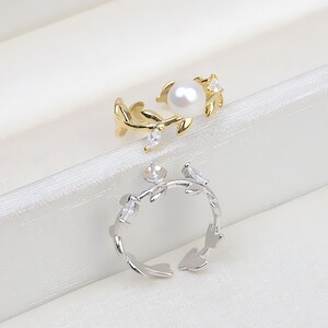 Può includere: Due anelli delicati con un design a foglia. Un anello è d'oro con una perla e pietre chiare, l'altro è d'argento con pietre chiare.