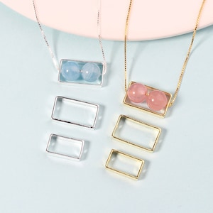 May include: Two necklaces with rectangular frames, one silver and one gold, each holding two round gemstones. The silver necklace has two smaller rectangular frames below it. The gemstones in the silver frame are light blue, and the gemstones in the gold frame are pink.