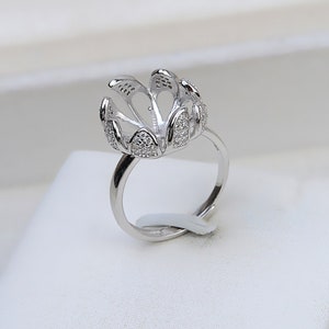 May include: A silver ring with a flower-shaped setting, designed to hold a pearl or other gemstone. The ring is adorned with small, clear stones.