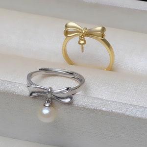 Könnte beinhalten: Zwei Ringe mit Schleifendesign. Der Ring links ist silber mit einem Perlenanhänger. Der Ring rechts ist gold mit einem kleinen Anhänger.