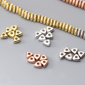 Puede incluir: Un conjunto de cuentas de forma triangular en oro, plata y oro rosa. Las cuentas tienen un pequeño agujero en el centro para enhebrar.