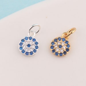 Könnte beinhalten: Zwei silberne und goldene Glücksbringer in Form eines bösen Auges mit blauen und weißen Steinen. Die Anhänger sind rund und haben oben eine Öse zum Anbringen an einer Halskette oder einem Armband.