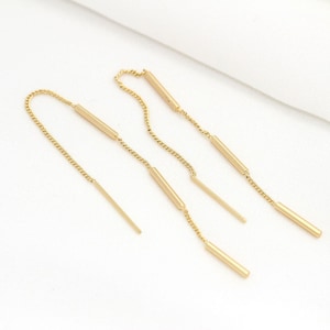 Könnte beinhalten: Ein Paar goldfarbene Kettenohrringe mit mehreren baumelnden Stäben. Die Ohrringe bestehen aus einer dünnen Kette mit kleinen, runden Gliedern. Jeder Ohrring hat drei Stäbe, die an der Kette baumeln. Die Stäbe haben alle die gleiche Länge und Breite und sind alle aus einem glatten, glänzenden Metall gefertigt.