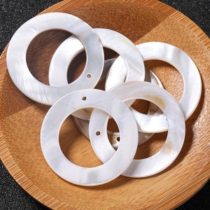 Op de afbeelding: Een groep van zes witte, ronde, schelpvormige ringen met een gat in het midden. De ringen zijn in een cirkel op een houten oppervlak gerangschikt.