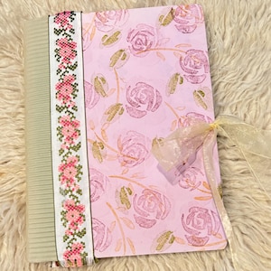 Pode incluir: Um diário feito à mão com uma capa estampada com rosas cor-de-rosa e uma fita floral decorativa. A lombada é de cor verde clara. O diário é amarrado com uma fita transparente amarelo claro.