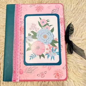 Puede incluir: Un diario floral rosa con lomo verde azulado y cierre de cinta negra. La portada presenta un diseño floral con un borde verde azulado y una ilustración central de flores azules y rosas. Un ribete de encaje rosa acentúa el lomo.