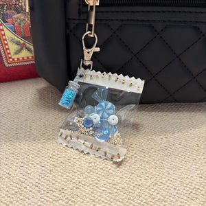 May include: A clear plastic bag charm filled with blue beads, candies, and glitter, attached to a black quilted backpack. The charm includes a small bottle of blue glitter and a variety of blue candies and beads.