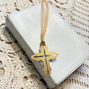 May include: A gold-colored cross bookmark with a decorative design hangs from a white book. The bookmark is attached to a thin, gold ribbon. The book has gold-colored text on the spine.