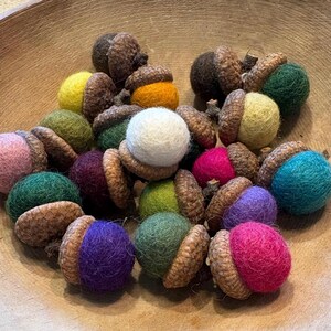 May include: A collection of decorative acorns featuring round, felted wool balls in various colors including pink, yellow, white, green, blue, and purple, each topped with a natural brown textured acorn cap.