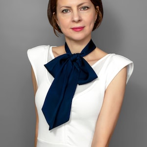 May include: A woman with short brown hair wearing a white sleeveless top with a large navy blue bow tie.
