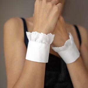 May include: A pair of white cotton wrist cuffs with lace trim. The cuffs are worn on both wrists and have a ruffled edge.
