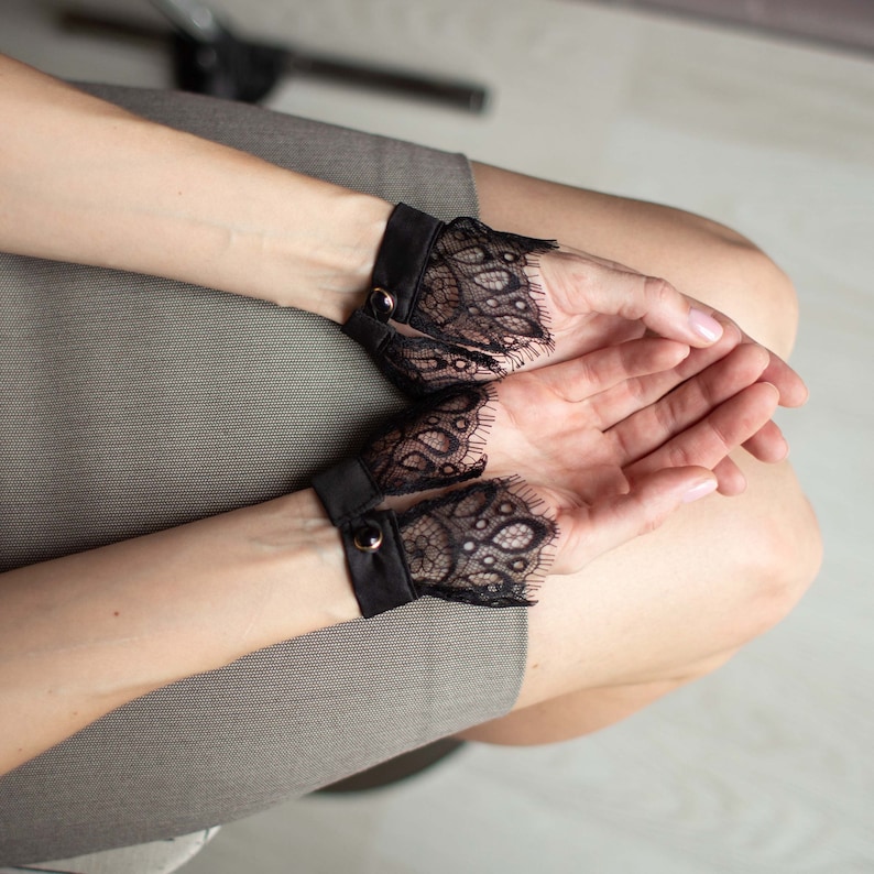 May include: Black lace wrist cuffs with a black band and gold button closure. The cuffs are worn on the wrists, with the intricate lace design extending over the hands. The background is a neutral gray.