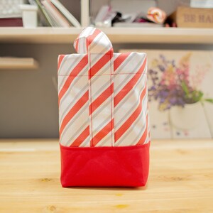 K&ouml;nnte beinhalten: Eine handgefertigte Geschenkt&uuml;te mit rotem Boden und einem K&ouml;rper mit diagonalen roten und grauen Streifen auf wei&szlig;em Hintergrund. Die Tasche hat einen passenden gestreiften Griff.
