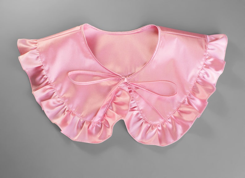 May include: A light pink satin collar with a ruffled edge and a bow tie closure. The collar is a soft, feminine accessory, perfect for adding a touch of elegance to any outfit. The collar is a delicate, romantic piece.